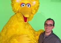 A man with glasses smiling next to the yellow Sesame Street character Big Bird against a green backdrop.
