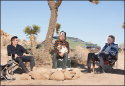 Three men sit in folding chairs around a rock fire pit in a desert with a car and Joshua trees.