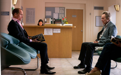 A man in a suit sits on a teal chair facing another man in a grey jacket in a waiting room.