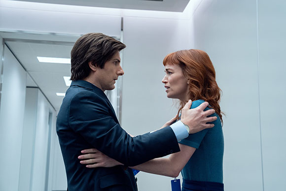 A man in a dark suit holds a woman with red hair by her shoulders in a bright hallway.