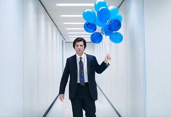 A man in a dark suit walks down a bright white hallway while holding a bunch of blue balloons.
