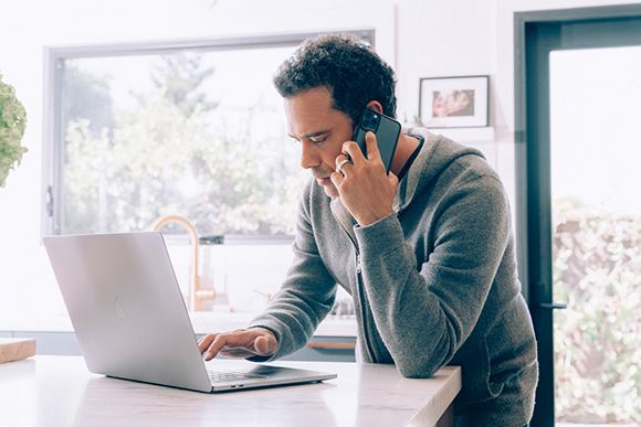 A man in a grey hoodie talks on a smartphone while leaning over a laptop on a kitchen counter.