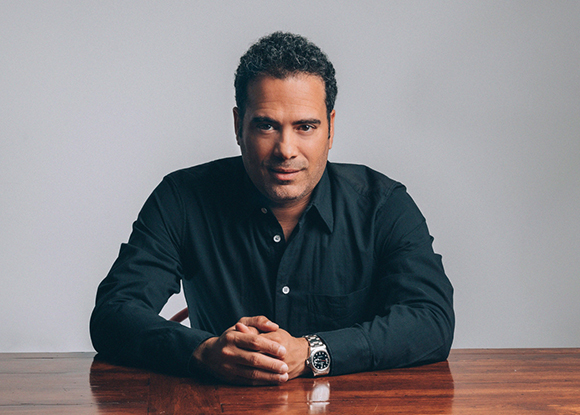 A man with dark curly hair wearing a black button-down shirt sits at a wooden table.