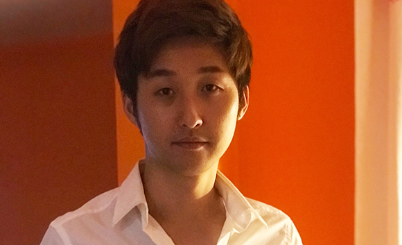A man with dark hair wearing a white collared shirt against an orange background.