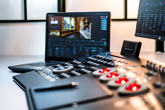 A laptop displaying video editing software next to a professional control surface with illuminated dials.