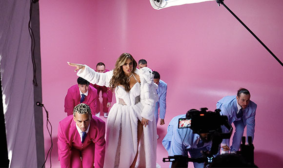 A woman in a white gown stands center stage among dancers in pink and blue suits against a pink backdrop.