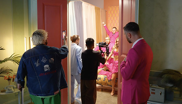 A film crew captures a scene of people in pink clothing through an open doorway.