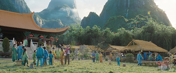 People in traditional blue and green clothing gather in a courtyard near a large temple and lush mountains.