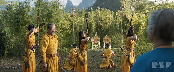 Archers in yellow traditional clothing aim bows in a lush green forest.