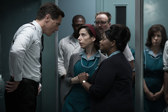 A man in a white shirt leans toward a woman in a teal uniform while others watch in a dim hallway.