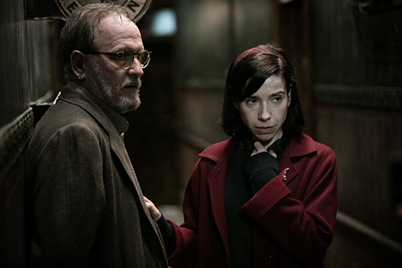 A man with glasses and a beard looks away while a woman in a red coat looks at him intensely.