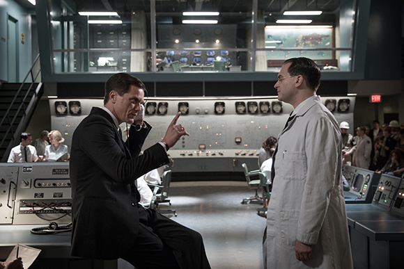 A man in a dark suit gestures toward a man in a white lab coat in a room filled with control panels.