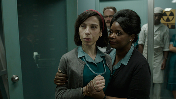 Two women in teal and grey medical uniforms stand close in a dimly lit hospital hallway.
