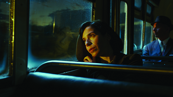 A woman gazes out a window of a bus at night, with a man sitting behind her.