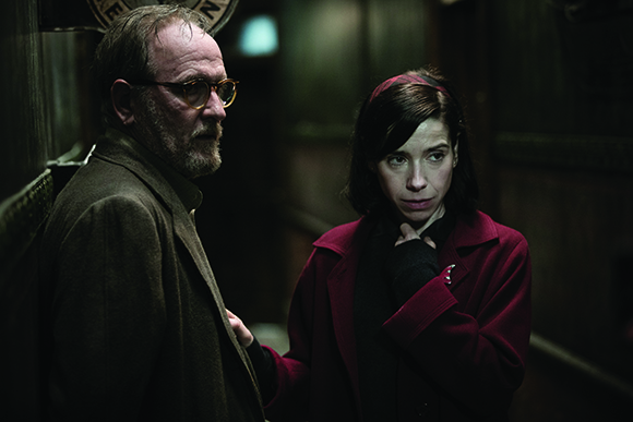 A man with glasses and a beard looks away while a woman in a red coat looks at him in a dark setting.
