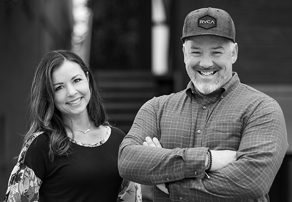 A smiling man in a baseball cap and plaid shirt stands with his arms crossed next to a smiling woman.