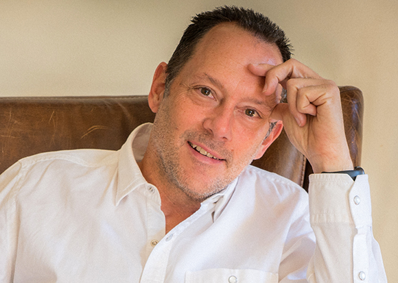 A man with short dark hair wearing a white button-down shirt, resting his head on his hand while sitting.