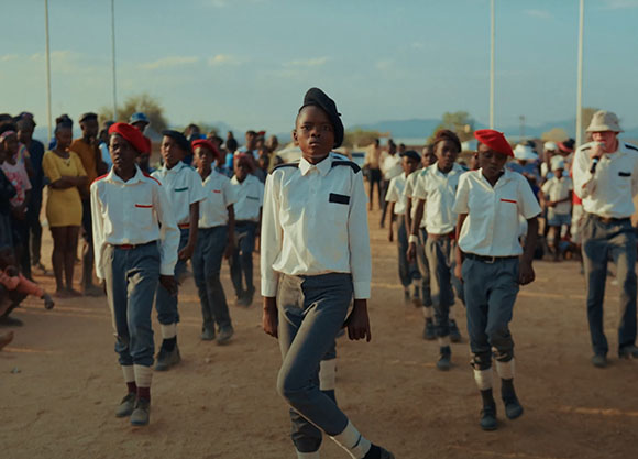 Students in white shirts and grey trousers march in formation on a dirt field under a clear sky.