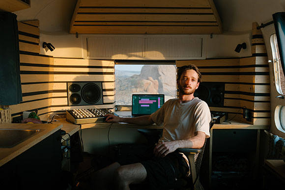 A man sits in a van converted into a music studio with speakers, a laptop, and a mixing console.