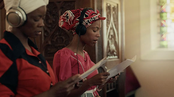 Two Black women wearing headphones and reading papers in a room with ornate wood carvings.