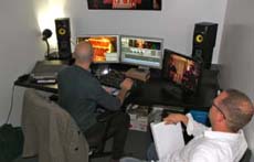 A man sits at a desk with three monitors and speakers, while another man watches from the foreground.