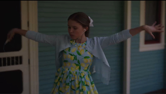 A young girl wearing a lemon-patterned dress and light blue cardigan dances with arms wide open.
