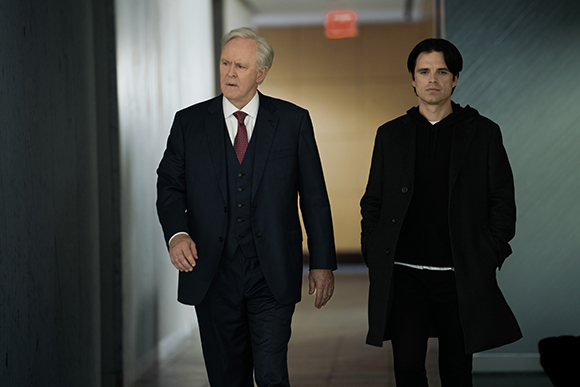 An older man in a dark suit and a younger man in a black coat walk down a hallway.