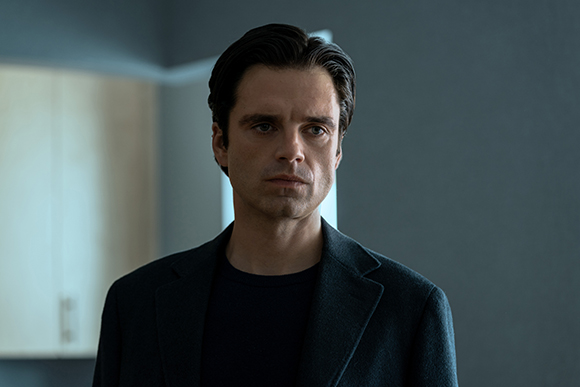 A man with dark hair and a serious expression wearing a dark coat and t-shirt.