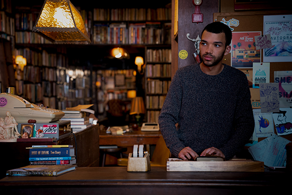 A man in a dark sweater stands behind a wooden counter in a dimly lit bookstore filled with books.