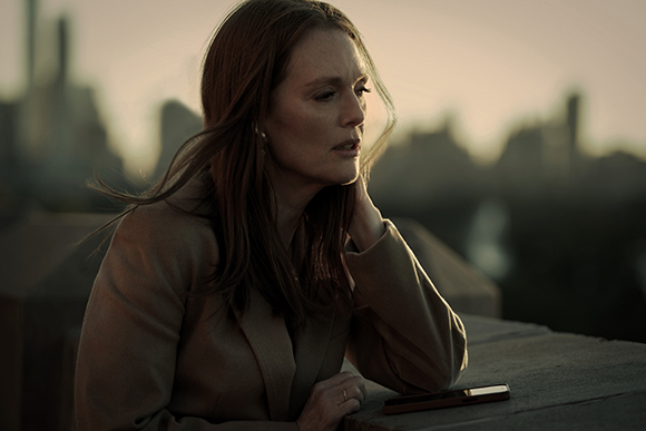 A woman with long brown hair leans on a stone ledge, looking away against a blurred city skyline at sunset.
