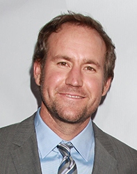 A man with short brown hair and a beard wearing a grey suit, light blue shirt, and striped tie.