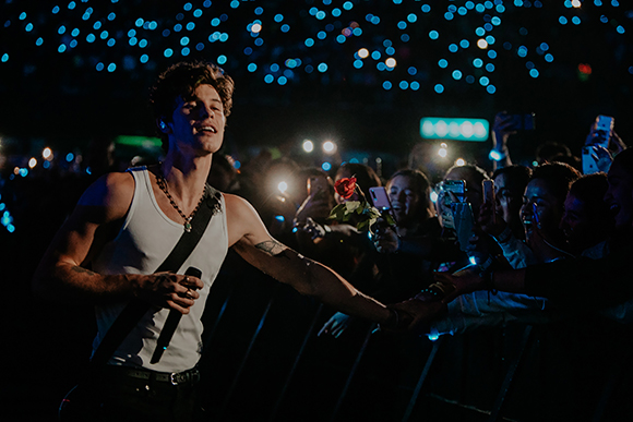 A male singer in a white tank top reaches out to touch hands with fans in a dark concert venue.