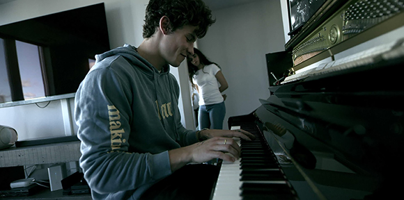 A young man in a grey hoodie playing the keys of a black piano in a room.