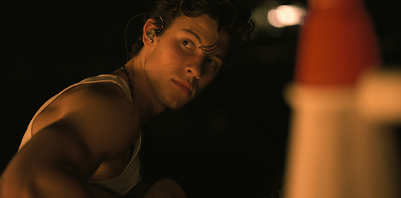 A young man wearing a white tank top and an earpiece looks upward against a dark background.