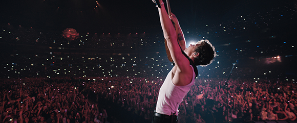 A man in a white tank top holds a guitar high above his head during a concert in a dark arena.