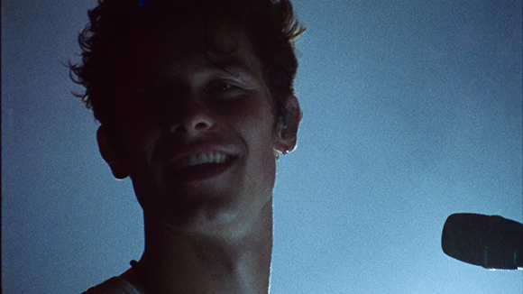 A low-angle close-up of a young man smiling under dim, blue-toned lighting.