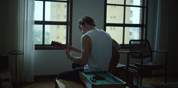 A person in a white sleeveless shirt sits by a window playing an acoustic guitar.