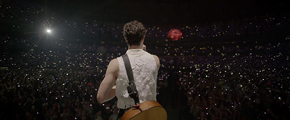 A musician seen from behind playing an acoustic guitar facing a large audience with glowing lights.
