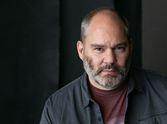A man with short hair and a grey beard looking directly at the camera against a dark background.