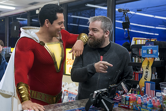 A man in a red and gold superhero costume smiles at a bearded man in a dark jacket behind a store counter.