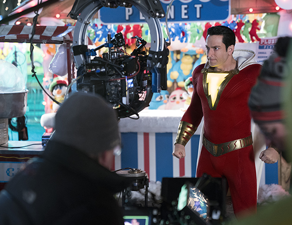 A man in a red and gold superhero costume stands on a film set behind a large movie camera.