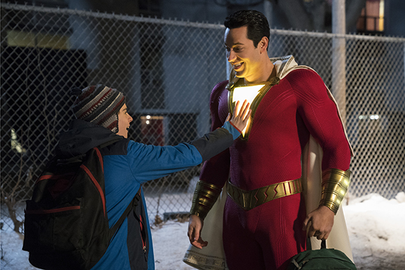 A boy in a blue jacket reaches out to touch the glowing yellow emblem on a man's red superhero suit.