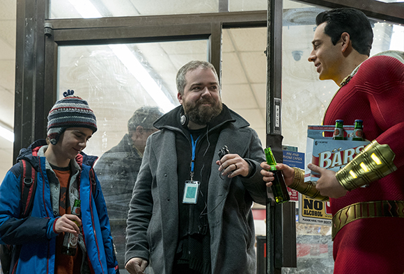 A man in a red superhero costume holds a six-pack of beer while talking to two men near a glass door.