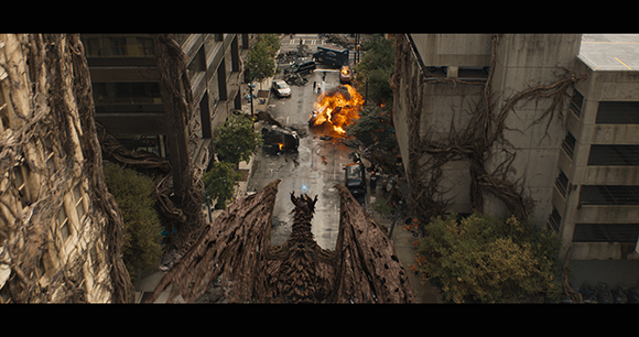 A large dragon flies over a city street with burning cars and vine-covered buildings.