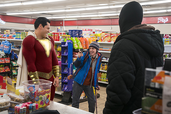 A man in a red and gold superhero costume smiles at a person filming him with a phone in a grocery store.