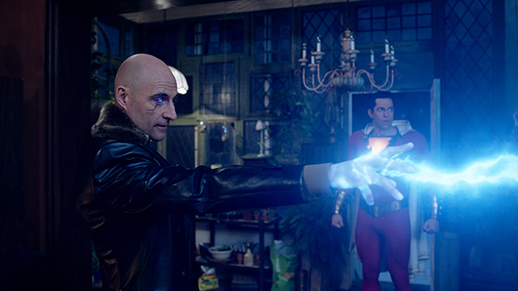 A bald man in a dark jacket reaches toward a bright blue bolt of energy in a dimly lit room.
