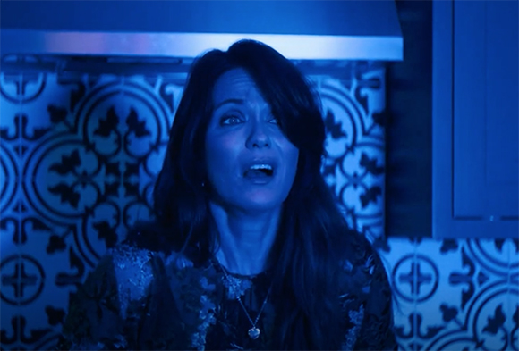 A woman with long dark hair looks upward with an open mouth in blue lighting.