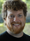 A smiling man with curly brown hair and a reddish-brown beard wearing a black shirt.