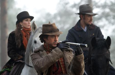 Three people in historical attire riding horses through a foggy, wooded area.