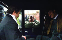 Three men in a dark vehicle; one in a suit sits left, one in a green jacket looks through a window, one smiles right.